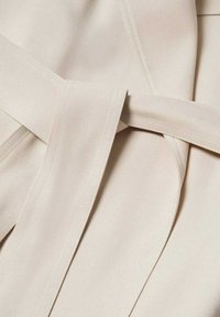 Beiger Stoff mit glatter Textur, ausgestattet mit einem gebundenen Taillendetail und einem breiten, flachen Gürtel. Sichtbare Nähakzente entlang der Kanten und Nähte.