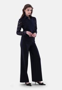 Marinebluse mit langen Spitzenärmeln, kombiniert mit hochgeschnittenen schwarzen, weiten Hosen und spitzen schwarzen Absatzschuhen mit Schleifenakzenten.