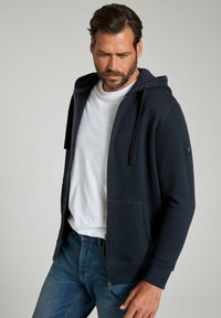 Marineblaues Zip-Hoodie mit einer Kapuze mit Kordelzug, Fronttaschen, gerippten Bündchen, aus strukturiertem Stoff, über einem weißen T-Shirt getragen.