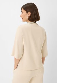 Beige, kortærmet top med en tekstureret overflade og en kontrasterende sort krave. Stile med matchende beige bukser, set bagfra.