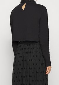 Svart kortärmad topp med långa ärmar, strukturerad tyg, som har en volangkrage och en nyckelhålsdetalj på baksidan, tillsammans med en svart kjol med stjärnmönster.