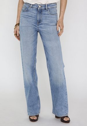 Jeans di un azzurro chiaro a gamba dritta indossati con una maglietta bianca, anelli e braccialetti dorati visibili, e sandali tacco aperti scuri.