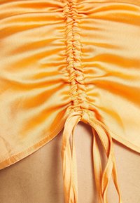 Detailní záběr na oranžovou, řasenou látku s uvázanými konci, která vykazuje hladkou texturu a zavěšený design, což naznačuje oděvní prvek.