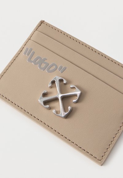 Θήκη καρτών από μπεζ δέρμα με τρεις θήκες για κάρτες, ασημένιο έμβλημα με διασταυρωμένα βέλη και αντανακλαστικό κείμενο "LOST" σε στυλιζαρισμένη γραμματοσειρά στην πάνω θήκη.