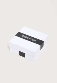 Λευκό τετράγωνο κουτί με μαύρη ταινία γύρω του, με το κείμενο "Calvin Klein" σε λευκά γράμματα πάνω στην ταινία.