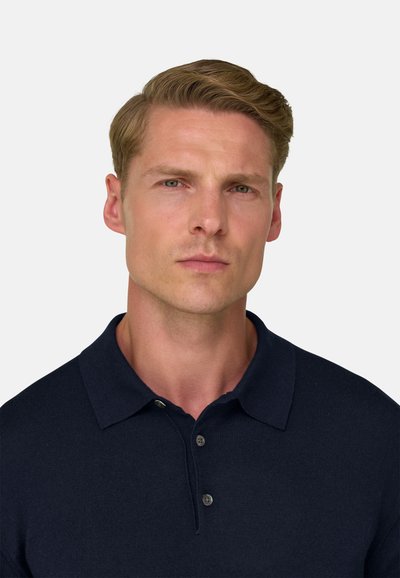 Homme blond aux cheveux courts portant une chemise bleu marine foncé à col, regardant droit devant avec une expression neutre sur un fond clair uni.