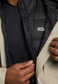Veste extérieure beige avec doudoune noire intérieure. Fermeture éclair partiellement ouverte, montrant la doublure noire et le logo de la marque. Mains tenant la fermeture éclair.