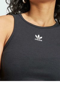 Kvinna som bär en svart ribbad ärmlös Adidas-topp med vit logotyp centrerad nära halsringningen och silverfärgade ringörhängen.