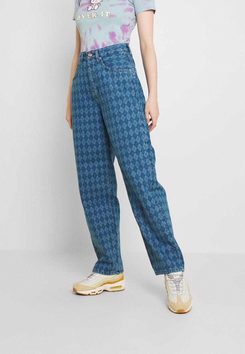 Osoba w niebieskich, wysokich dżinsach we wzór diamentów, fioletowej bluzie tie-dye z nadrukiem kreskówkowym oraz w białych i beżowych trampkach stoi na gładkiej podłodze.