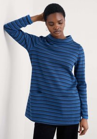 Kobieta z krótkimi włosami, ubrana w niebiesko-czarno paskowaną koszulę z długim rękawem, stojąca na tle białej ściany z uniesioną jedną ręką za głowę.