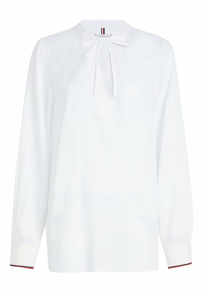 Blusa bianca con maniche lunghe, un collo rotondo e un dettaglio a fiocco sul davanti. Presenta polsini a contrasto con un accento di colore. Tessuto morbido.