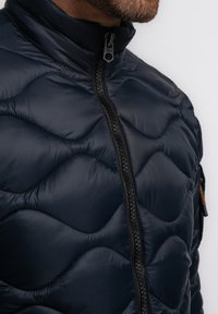 Marineblaue, knuffige Jacke mit hohem Kragen, ausgestattet mit einem strukturierten, gesteppten Design und einem schwarzen Frontreißverschluss, mit einem kleinen braunen Akzent.