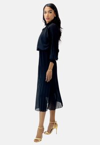 Vestido midi negro plisado con mangas largas abullonadas y un corpiño ajustado. Acentuado con unos zapatos de tacón alto dorados con tiras. Tejido ligero y suave.
