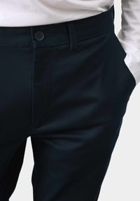 Marineblaue Hose aus glattem Stoff, mit einem einzelnen Knopfverschluss, Gürtelschlaufen und seitlichen Taschen für Funktionalität.