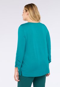Žena s blond vlasy, oblečená v tyrkysovém dlouhém rukávu s řasenými rukávy a odpovídajícími kalhotami, zobrazená zezadu na bílém pozadí.