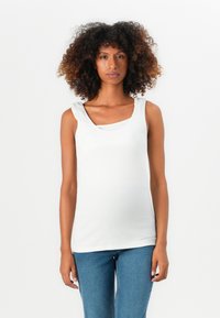 Ung kvinne med krøllete hår iført en hvit ermeløs topp og blå jeans, stående mot en enkel hvit bakgrunn.