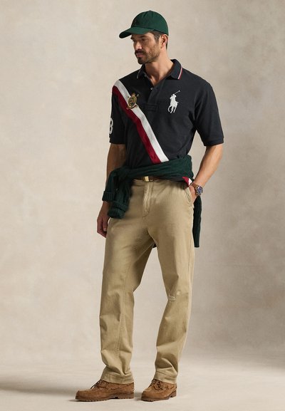 Mann som har på seg svart poloskjorte med hvite og røde diagonale striper, beige bukser, grønn caps, grønn genser knyttet rundt livet og brune sko.