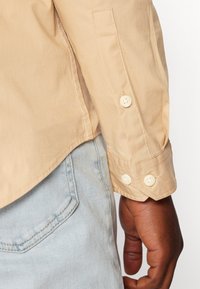 Camicia beige con bottoni e maniche arrotolate e bottoni bianchi. Jeans azzurri chiari visibili nella parte inferiore dell'immagine. Texture del tessuto liscia.
