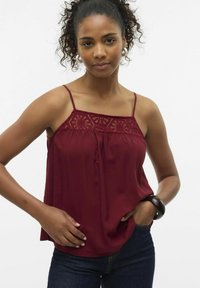 Bordeaux mouwloos topje met een kanten geborduurd lijfje, losse pasvorm en dunne schouderbandjes, gecombineerd met denim jeans en een donkere armband.