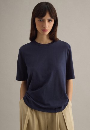 Giovane donna con capelli castani lisci che indossa una maglietta a maniche corte blu navy e pantaloni beige plissettati su uno sfondo neutro.