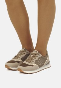 Zapatillas beige y marrón con materiales de malla y ante, que cuentan con cierre de cordones, detalle del logo y suelas de goma. Diseño de perfil bajo.