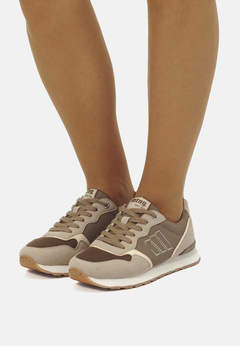 Zapatillas beige y marrón con materiales de malla y ante, que cuentan con cierre de cordones, detalle del logo y suelas de goma. Diseño de perfil bajo.
