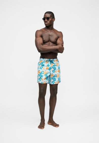 Μυώδης άνδρας που φοράει μαύρα γυαλιά ηλίου και πολύχρωμα μαγιό με λουλούδια στέκεται ξυπόλητος με σταυρωμένα τα χέρια μπροστά σε απλό φόντο.