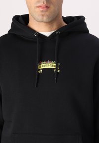 Black hoodie featuring a front graphic with yellow banner text "SANTA CRUZ" and floral accents. Made of soft, thick fabric with a kangaroo pocket.