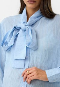 Lichtblauwe blouse met een grote strik bij de hals, met een gekreukeld textuur en een knoopsluiting aan de voorkant. Zachte stof en manchetten zijn zichtbaar.
