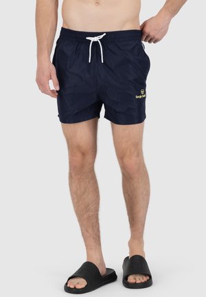 Shorts de bain navy avec une taille à cordon de serrage, arborant un logo jaune sur la cuisse gauche et un tissu léger et texturé. Portés avec des tongs noires.