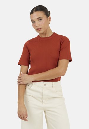 Vrouw met donker haar in een knot, gekleed in een roestkleurig T-shirt met korte mouwen en crèmekleurige broek met hoge taille, staand met één arm over de andere gekruist.