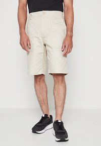 Hombre vistiendo pantalones cortos beige hasta la rodilla, zapatillas negras y una camiseta negra, de pie frente a un fondo blanco liso.