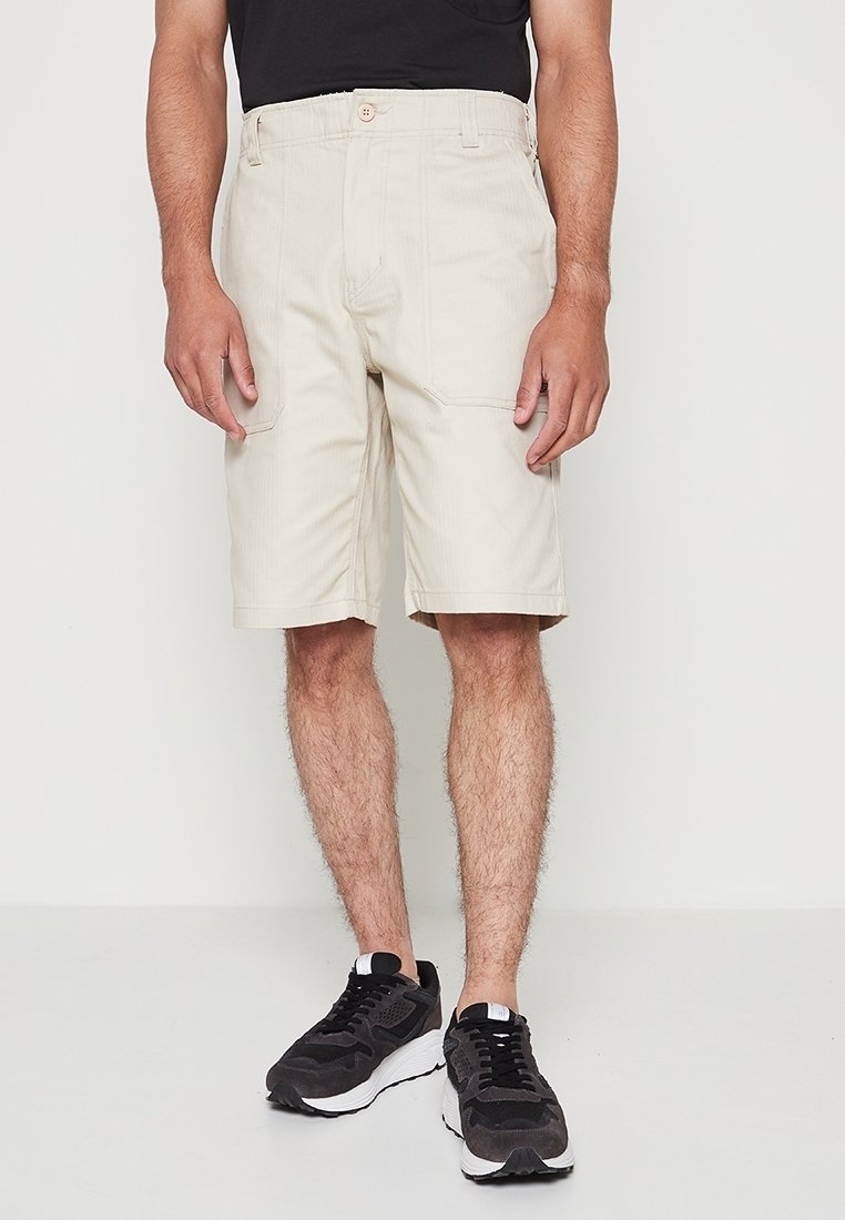 Hombre vistiendo pantalones cortos beige hasta la rodilla, zapatillas negras y una camiseta negra, de pie frente a un fondo blanco liso.