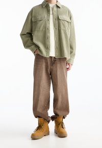 Person, der ein hellgrünes Cordhemd, ein beiges T-Shirt, lockere braune Hosen und sandfarbene Schnürstiefel vor einem weißen Hintergrund trägt.