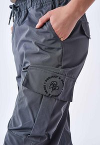 Pantaloni cargo grigi con vita elasticizzata, tasche laterali e una patch con la scritta "Project X Paris". Texture liscia, tessuto resistente.