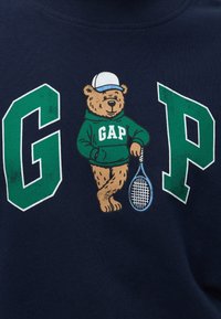 Ours portant un sweat à capuche GAP vert et une casquette blanche, appuyé sur une raquette de tennis entre les grandes lettres vertes G et P sur un tissu foncé.
