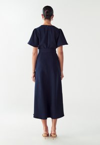 Námorníckomodrá šaty s krátkymi nafúknutými rukávmi, obtiahnutým pásom a dĺžkou po členky. Hladká látka so zipsom na zadnej strane.