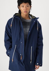 Marineblauwe parka-jack met een capuchon, voorzien van een rits aan de voorkant, trekkoordaccenten en twee voorkruiszakken. Gemaakt van een glad, duurzaam materiaal.