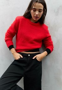 Maglione rosso, con texture, a girocollo e polsini neri, abbinato a pantaloni neri a vita alta con bottone frontale e tasche.