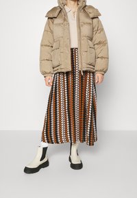 Beige puffervest med stor krave, knaplukning og to forlommer. Undervesten en mønstret maxiskirt i brune og creme cirkler. Sort og hvide ankelstøvler.