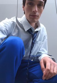 Vīrietis ar gaiši zilu krekli un zilu un baltu svītrainu kaklasaiti. Viņam ir zilas bikses un neitrāla fona. Fokusēts uz augšējo ķermeņa daļu un seju.