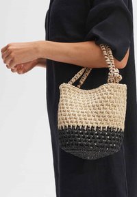 Geweven mand handtas met een natuurlijk beige en zwart ontwerp, gestructureerde handvatten en een ronde vorm, vastgehouden door een hand.