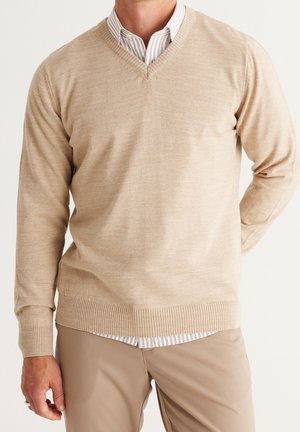 Hombre vestido con jersey beige de cuello en V sobre camisa blanca de rayas y pantalones beige, de pie con una mano detrás de la espalda.