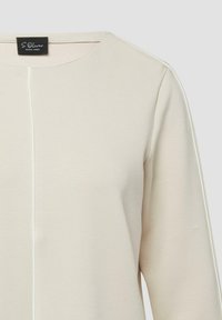 Haut beige à manches longues avec un col rond, présentant une couture centrale et de subtiles touches blanches sur les épaules.