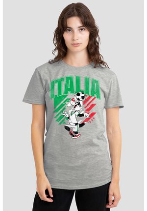 Junge Frau trägt ein graues T-Shirt mit grünem "ITALIA" Text, den Farben der italienischen Flagge und einem Cartoon-Hund, der Fußball spielt und einen Ball auf dem Kopf balanciert.