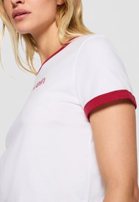 White t-shirt with a round neckline, short sleeves featuring burgundy trim, and embroidered detailing on the chest. Soft cotton material.