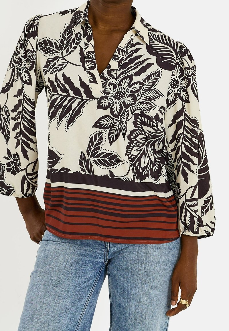 Blouse florale noire et blanche à manches trois-quarts avec des rayures rouges et noires à l'ourlet, portée avec un jean bleu clair.