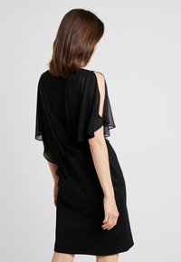 Robe noire avec manches évasées transparentes, une silhouette ajustée et une texture lisse, mettant en valeur un design à épaules dénudées et une coupe longueur genou.
