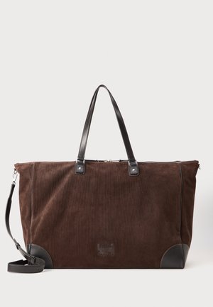Borsa duffel in velluto a coste marrone con accenti in pelle nera, manici doppi, tracolla regolabile e chiusura con zip. Presenta una superficie texturizzata e un logo.