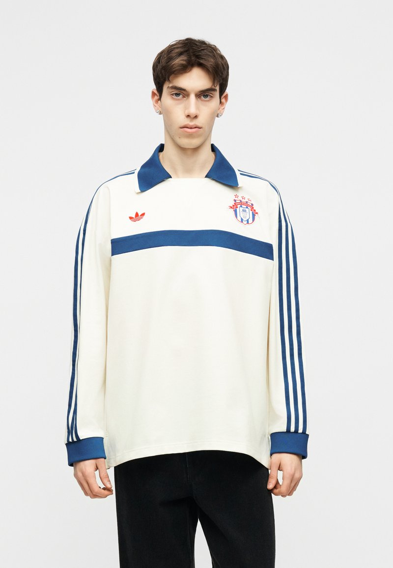Giovane uomo che indossa una maglia Adidas bianca a maniche lunghe con strisce blu sulle maniche, colletto blu e logo ed emblema rossi sul petto.
